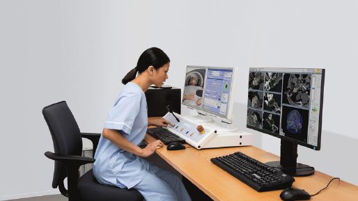 Photograph of a female health care worker speaking to a Elekta Leksell Gamma Knife icon patient via control console. There are two monitors, one showing the camera feed and dashboard, and another showing scan results.